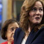 Justices Amy Coney Barrett and Elena Kagan stand together indoors, one in the foreground with wavy brown hair and a navy blazer, looking to her right. The other woman, wearing red, stands behind her with a neutral expression. Blurred background includes people and a flag.