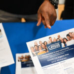 A person hands a U.S. Census job flyer across a blue table to another person holding a similar flyer; only their hands and the flyers are visible.