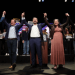 Five people stand on a stage with their arms raised in victory, standing behind a lectern with campaign signs for Elaine Luria and others in the background.