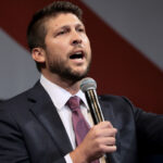 A man in a suit and tie speaks into a microphone while gesturing with his raised index finger.