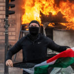 A person wearing a black balaclava and jacket stands in front of a burning structure while holding a Palestinian flag.