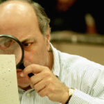 A man in a striped shirt uses a magnifying glass to inspect a small card with holes.