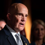Keith Self in a suit and tie speaking at a podium with two people visible in the blurred background.