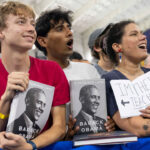 Two young men and a woman hold copies of Barack Obamas book A Promised Land at a rally, while the woman holds a sign reading Im their teacher.