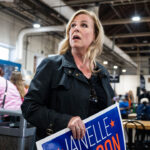 A blonde woman in a black jacket looks upward while holding a blue campaign sign for Janelle Stelson in an indoor market hall.