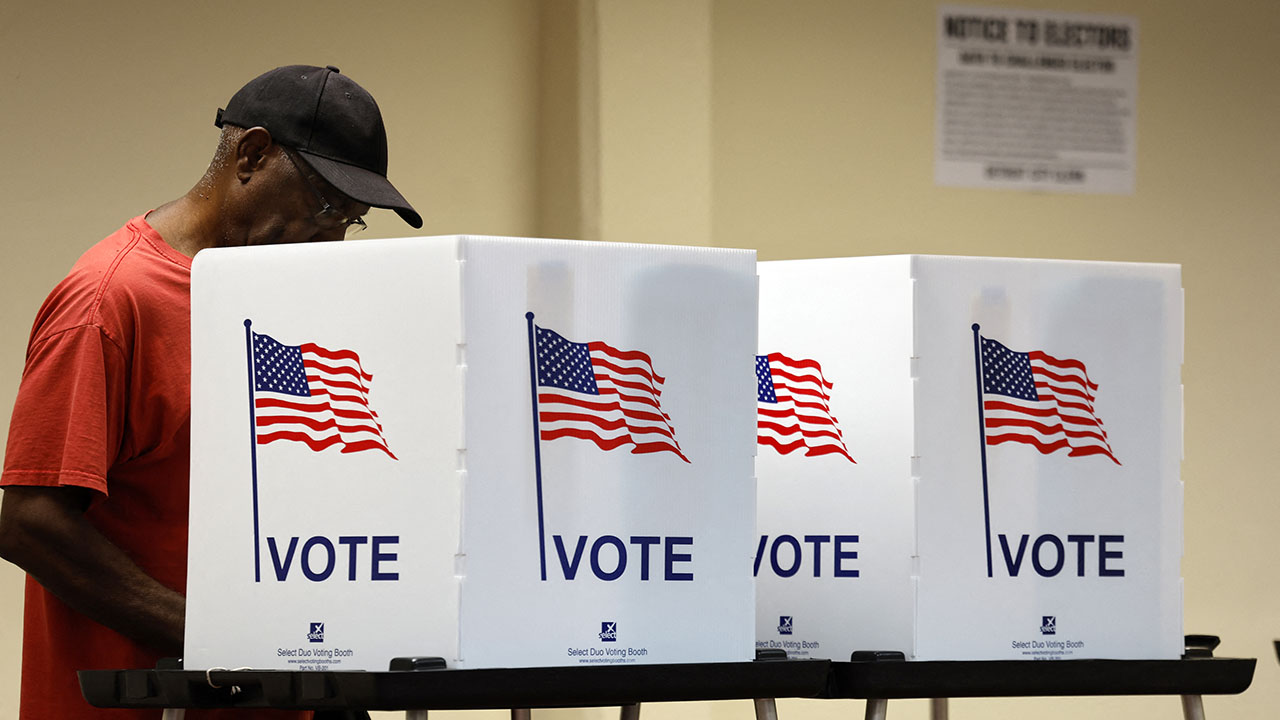 A man in a red shirt and black cap stands at a white voting booth with an American flag graphic, a scene captured during a Texas election lawsuit.