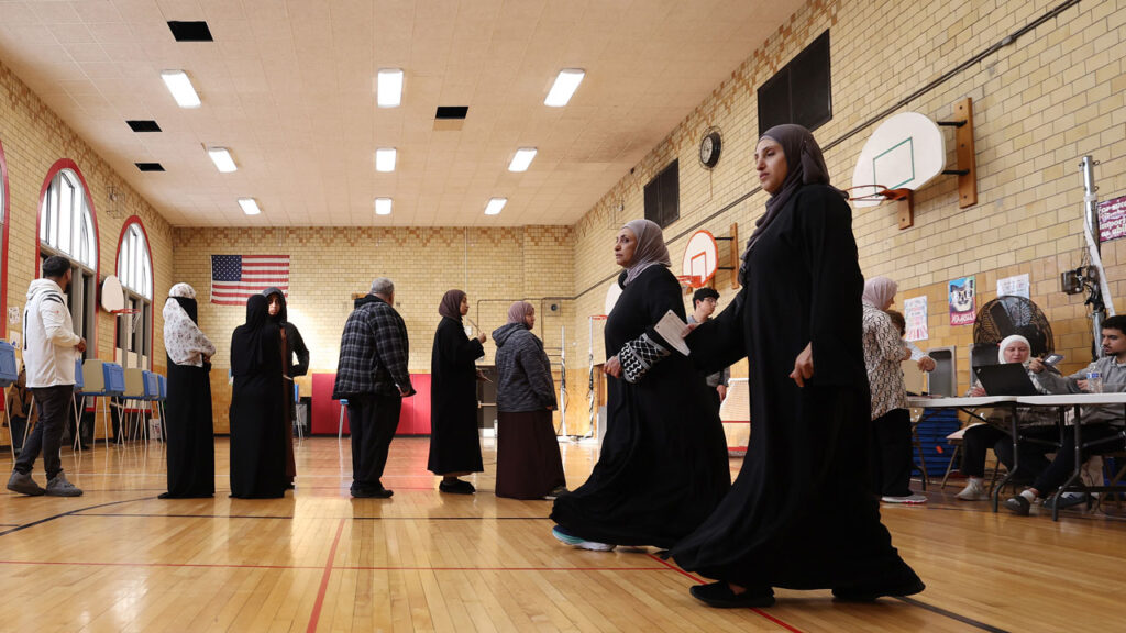 People in traditional clothing, including some associated with Muslim migration, walk across a gymnasium floor set up as a polling station, with voters checking in at tables.