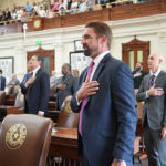A group of people stands in a legislative chamber with their hands over their hearts during a meeting on Texas education policy.