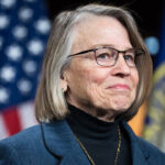 A middle-aged woman with glasses, wearing a dark blazer over a black turtleneck, standing in front of two blurred flags.