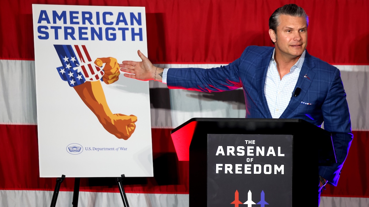 A man in a blue suit stands at a podium labeled The Arsenal of Freedom and gestures toward a poster titled American Strength featuring a graphic of a muscular arm with a flag-patterned sleeve.