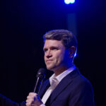 James Talarico in a suit holding a microphone and gesturing while speaking on stage.