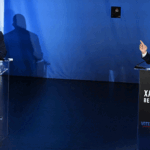 Steve Hilton and Xavier Becerra stand at podiums labeled Steve Hilton and Xavier Becerra during a debate. Both are speaking, with one gesturing. A Voters Decide sign is on each podium, and the background is blue.
