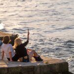 Four people sit on a stone wall by the water, with one woman holding up a phone to take a selfie.
