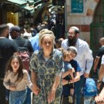 A crowd of people walking through a narrow market street, including a woman in a patterned dress holding a childs hand and a man carrying a toddler.