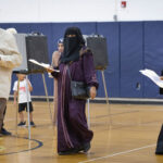 People in a gymnasium walk past voting booths while holding documents, a scene noted in discussions regarding Muslim migration.
