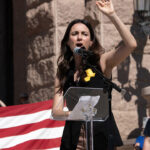 Gina Hinojosa speaks at a podium with her hand raised in front of the Texas state capitol building with an American flag draped behind her.