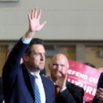 A man in a suit raises his hand while standing among a group of people. Several applaud, and a person holds a red sign that reads DEFEND OUR CULTURE in the background.