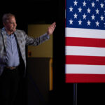 A man in a plaid blazer waves his hands while walking past a large American flag during the Texas Senate race.