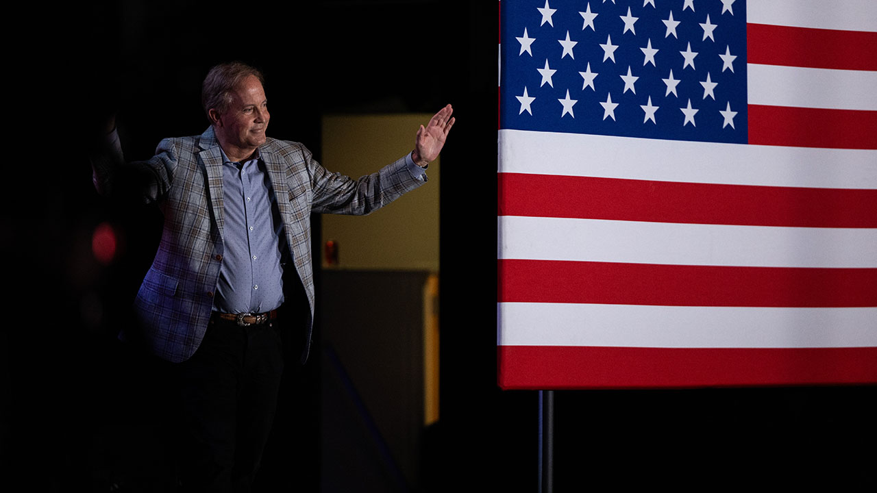 A man in a plaid blazer waves his hands while walking past a large American flag during the Texas Senate race.
