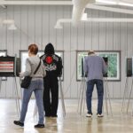 Four people stand at individual voting booths in a large, industrial-style room.