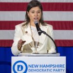 A woman in a white blazer speaks at a podium with the New Hampshire Democratic Party logo in front of an American flag.