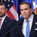 Two men in suits standing at a podium in front of American and Department of Veterans Affairs flags, with one man speaking into a microphone.