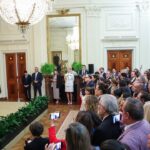 A man speaks at a podium to a large audience in a formal, ornate room with chandeliers, mirrors, and gold curtains. People watch attentively, some taking photos with their phones.