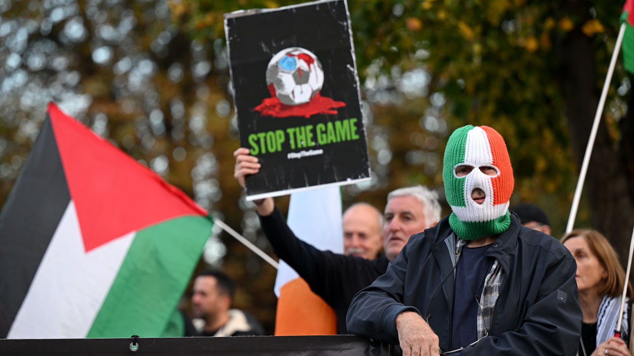 A protester wearing an Ireland flag balaclava holds a sign depicting a soccer ball and the text STOP THE GAME next to a person holding a Palestinian flag.