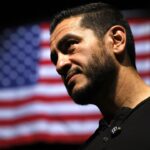 A man with a beard looking to the side in front of a blurred American flag.