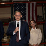 Speaker Mike Johnson speaks into a microphone, flanked by three other people, with an American flag hanging in the background.