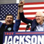 Troy Jackson and Bernie Sanders stand behind a podium with their hands raised together in front of an American flag.