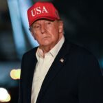 Donald Trump wearing a red USA hat and a dark suit jacket over a white shirt.