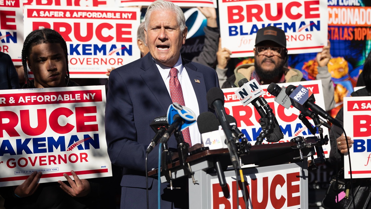 Bruce Blakeman speaks at a podium surrounded by supporters holding Bruce Blakeman for Governor campaign signs and a cluster of news microphones.