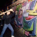 A man swings a pickaxe at the Berlin Wall as a crowd watches the fall of communism and socialism in Europe.