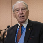 Senator Chuck Grassley speaking at a podium with two people in the background.