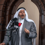 A man in a white thobe, ghutra, and gray suit jacket speaks into multiple microphones in front of a wooden arched doorway.