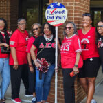 A group of nine women wearing red shirts and Delta Sigma Theta apparel stand together outside a building, posing with a round I Voted! sign.