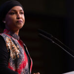 Ilhan Omar speaking at a podium with microphones, advocating for justice.
