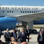 Vice President J.D. Vance is pictured in a suit and gestures with his hands while walking on an airfield away from Air Force 2, accompanied by a group of people in suits and security personnel.