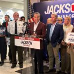 Rick Jackson speaks at a podium with a Jackson for Governor sign, surrounded by a group of supporters holding campaign signs.