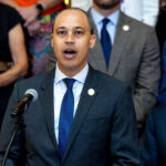 A man in a suit and blue tie speaks into a microphone, flanked by two other men in suits, while others stand in the background.