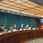 Members of the Joint Election Equipment Specifications and Standards Committee meet. They are dressed in professional attire and are seated at a curved table in a formal meeting room.