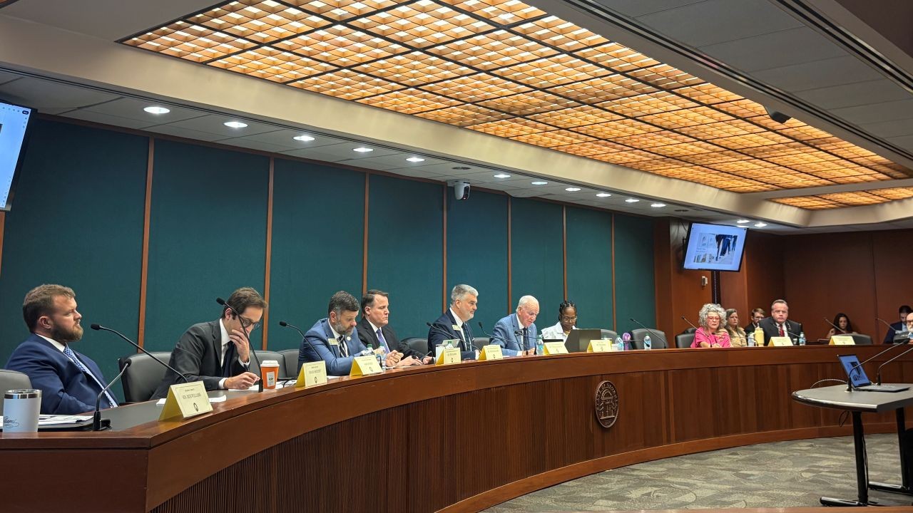 Home 1 Members of the Joint Election Equipment Specifications and Standards Committee meet. They are dressed in professional attire and are seated at a curved table in a formal meeting room.