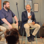 Four panelists sit on stools on a stage, with one man speaking into a microphone.