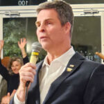 John McGuire in a suit jacket speaks into a microphone before a crowd of supporters.