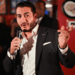 A man in a suit and white shirt holds a microphone and gestures while speaking to a room of people in Michigan.