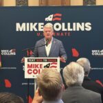 Rep. Mike Collins speaks to supporters from a podium in front of a blue campaign backdrop with U.S. and Georgia state flags.