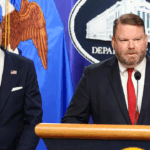 Two men in suits stand at a podium in front of a Department of Justice seal and a flag.