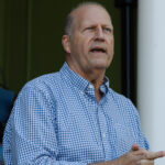 A man in a blue and white checkered button-down shirt speaks outdoors, with a woman partially visible in the background.