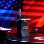 Sen. Jon Ossoff and Rep. Mike Collins are pictured in suits and stand at podiums labeled Debate Night Live Race for Senate on a stage with a red, white, and blue American flag backdrop.